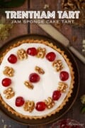 A whole jam sponge cake tart with icing, cherries and walnuts with text overlay.