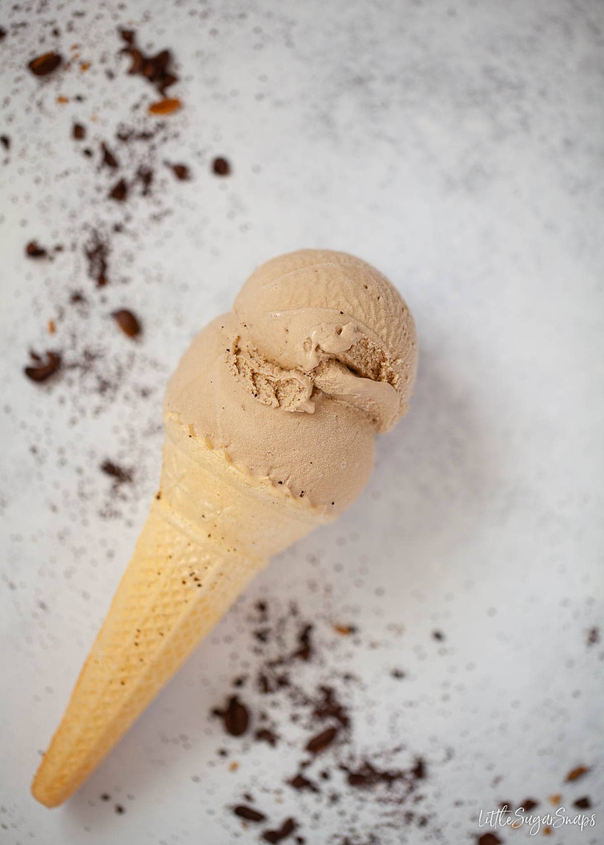 A double scoop of coffee ice cream in a wafer cone with ground coffee scattered around.