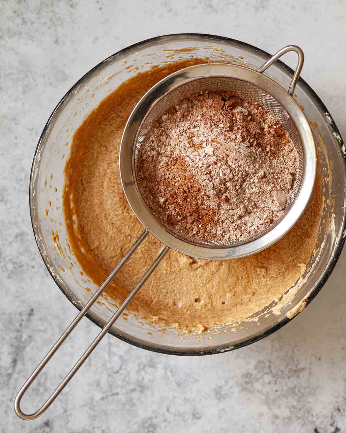 A sieve holding flour, spices and cocoa powder suspended over a bowl of cake batter.