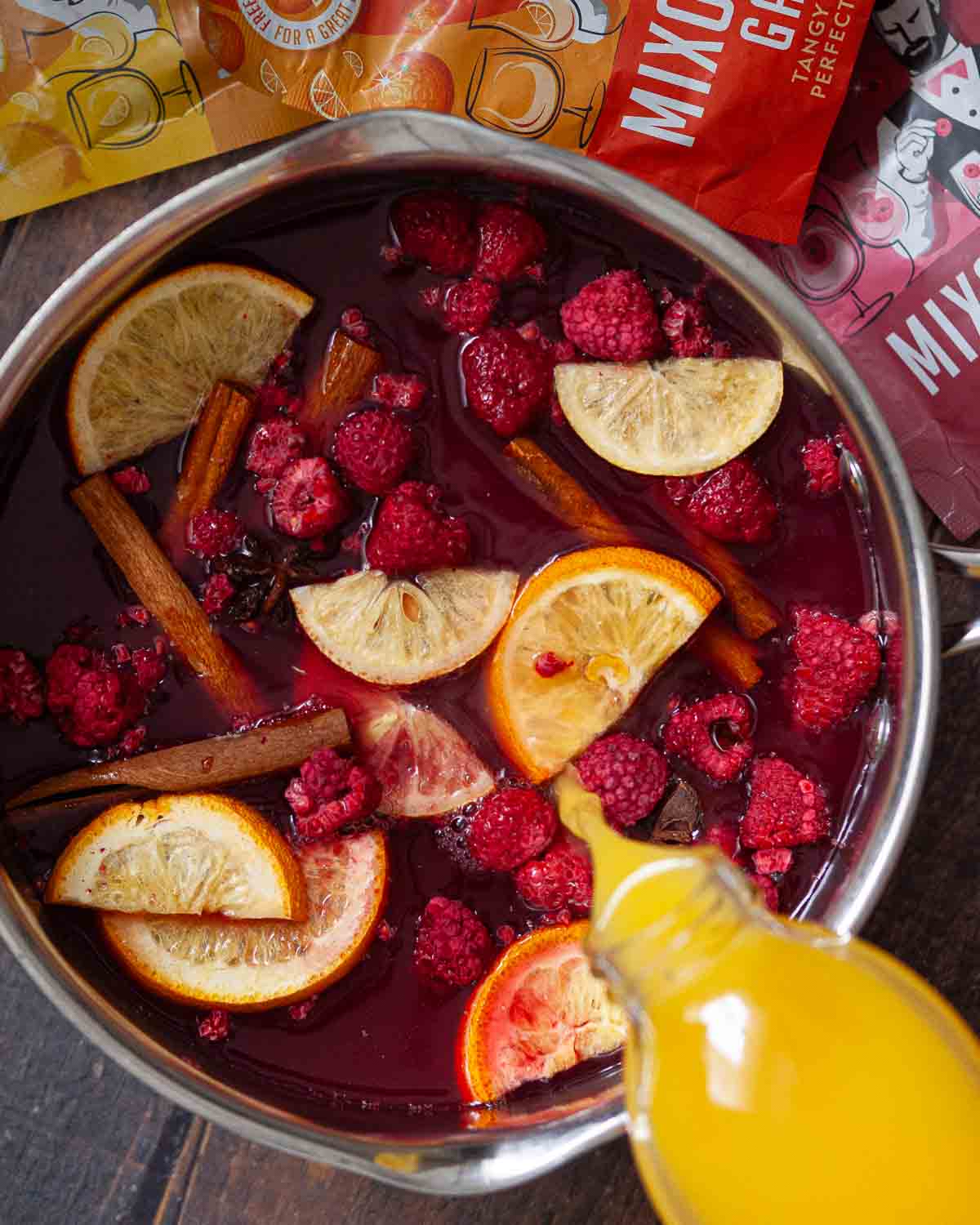Orange juice being poured into a pan with other juice, spices, lemons, oranges and raspberries.
