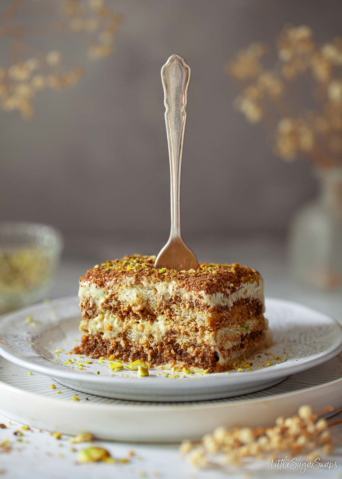 A slice of coffee and pistachio cream dessert on a plate with a fork standing up in it.