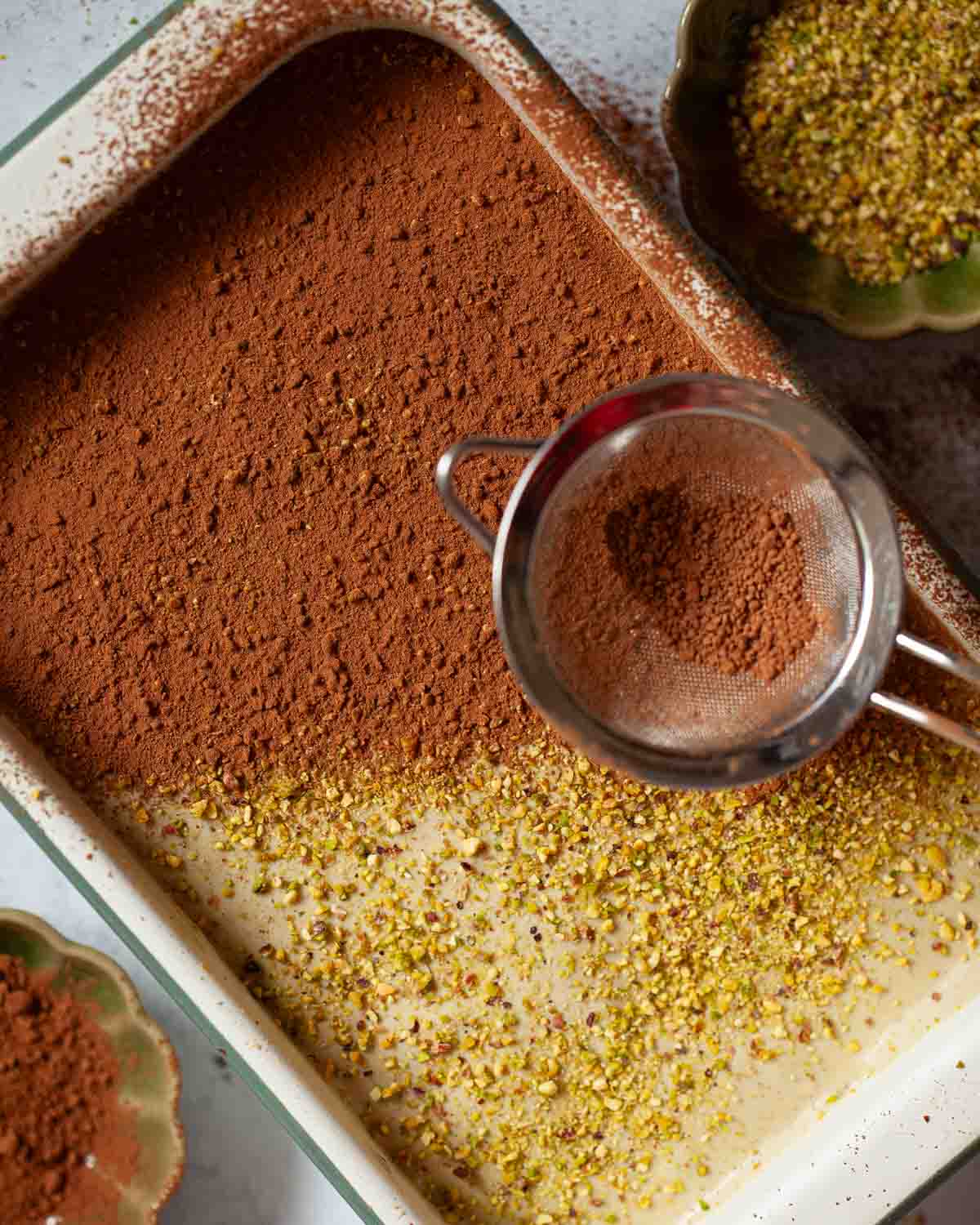 Cocoa powder being sieved over pistachio tiramisu.