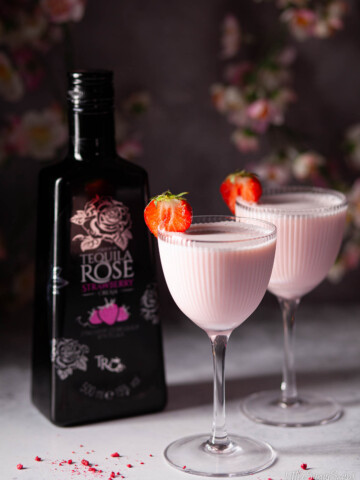 Two pink creamy strawberry liqueur cocktails next to a bottle of strawberry cream liqueur.
