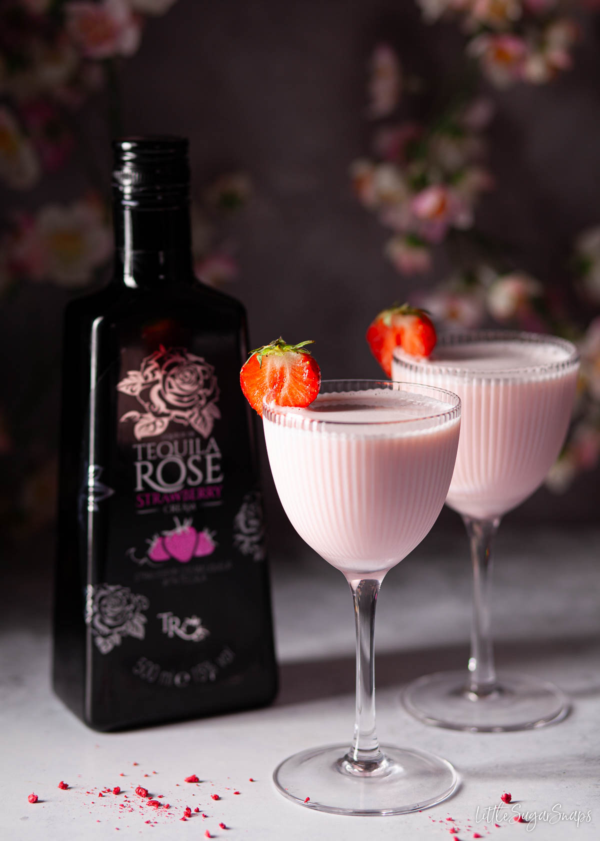 Two pink creamy strawberry liqueur cocktails next to a bottle of strawberry cream liqueur.