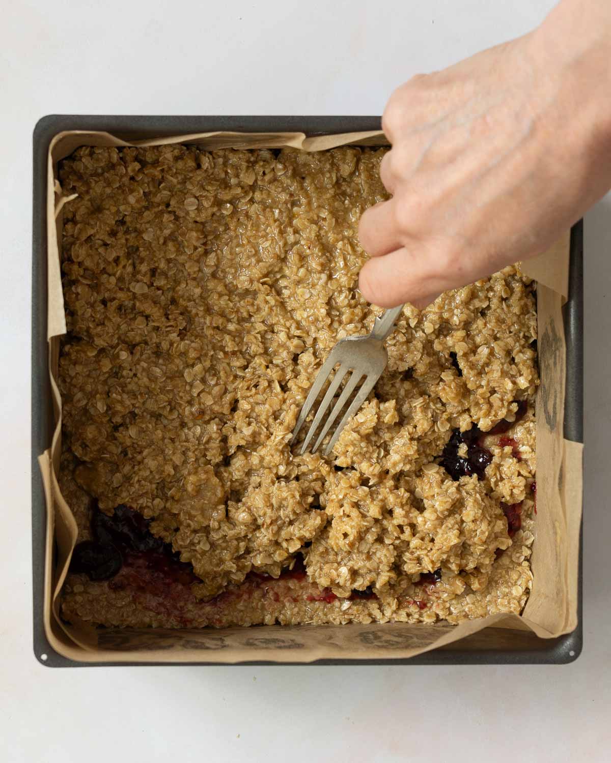 A person spreading flapjack mix over a layer of jam in a baking tin.