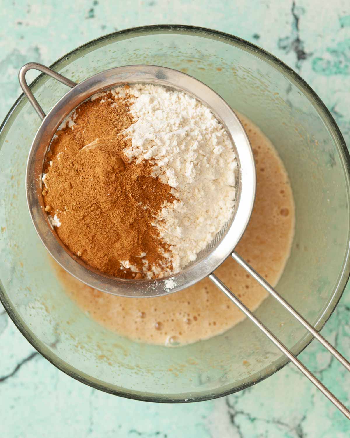 Flour and baking spices in a sieve suspended over a bowl of cake batter.