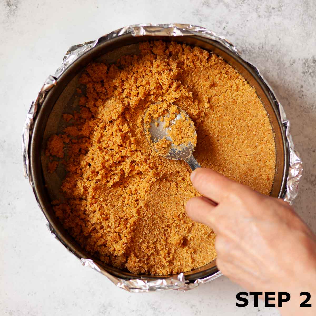 A person pressing cheesecake biscuit base into a baking tin that is wrapped in foil.