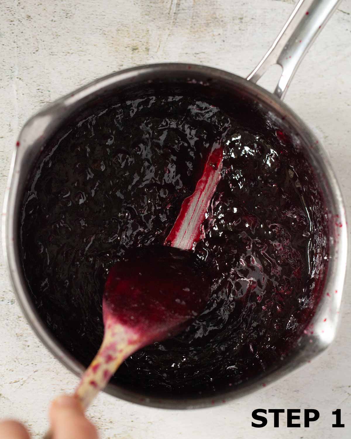 a person running a spoon through blackcurrant compote in a pan.