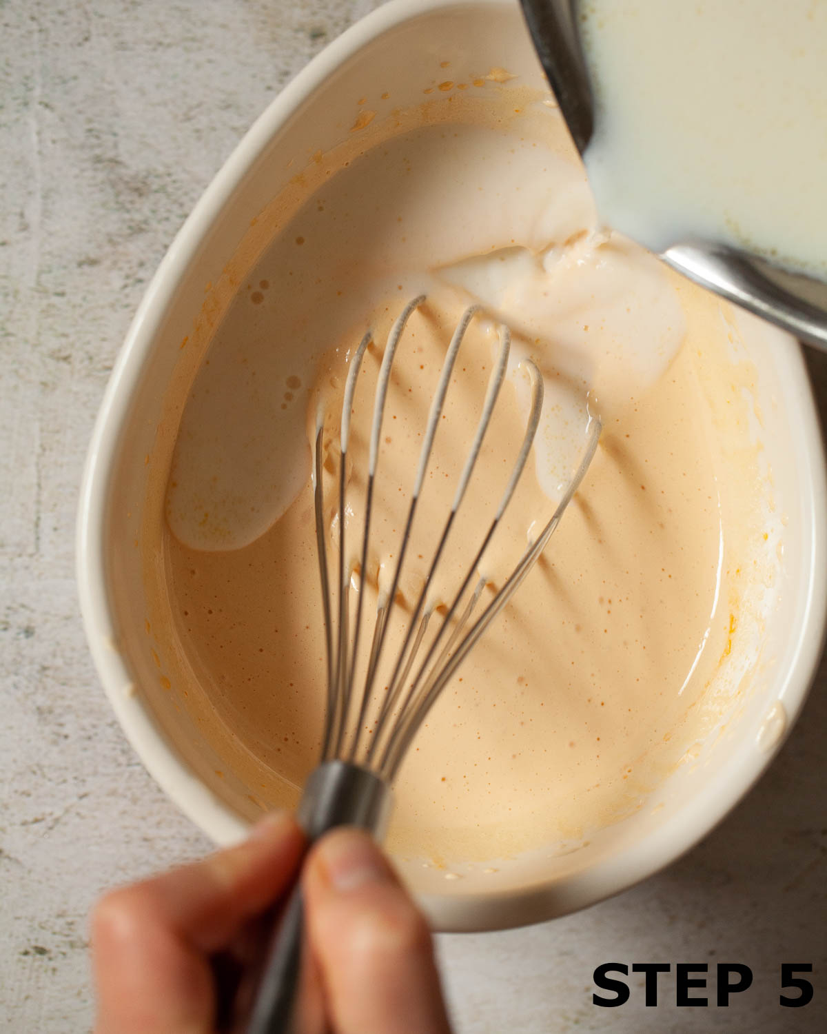 A person pouring hot milk into whisked egg yolks and sugar for custard.