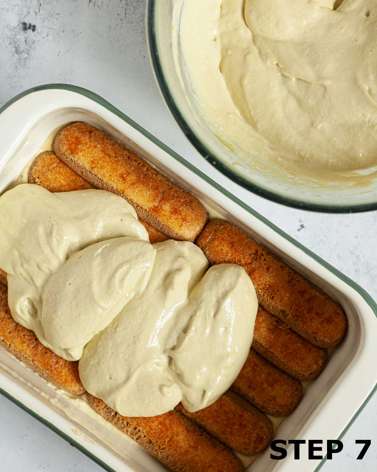 A layer of coffee-soaked ladyfingers being topped with pistachio cream in a serving dish.