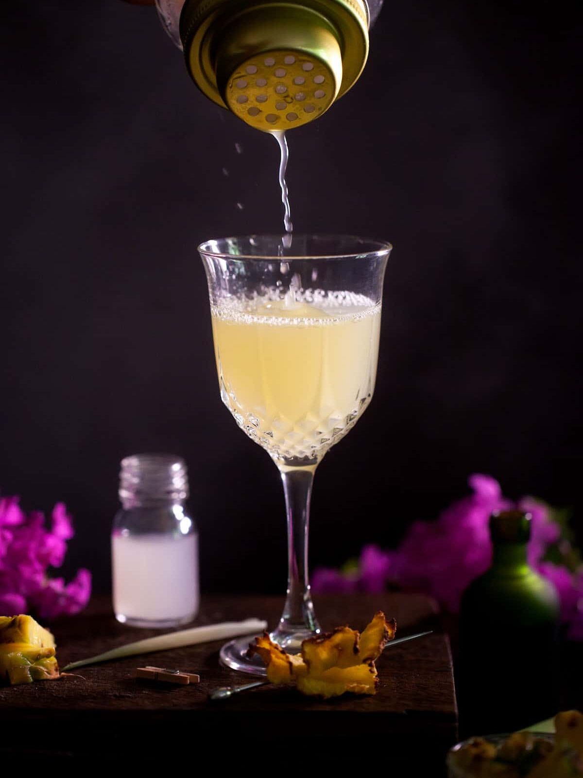 Pouring a royal hawaiian cocktail into a nick and nora style glass. A small jar of orgeat syrup and purple flowers are out of focus behind the glass