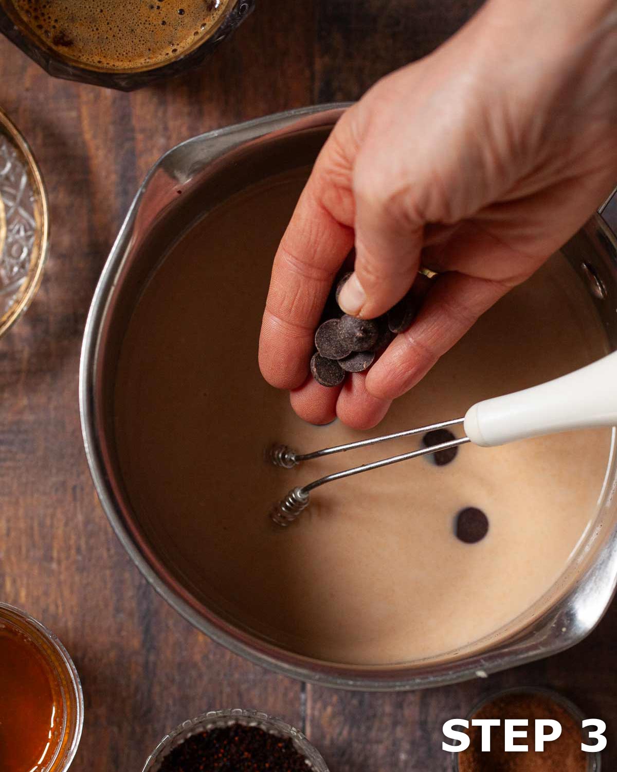 A person adding dark chocolate chips to spice-infused milk in a pan.