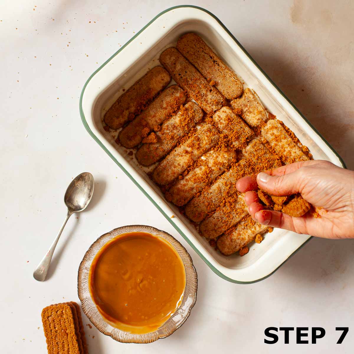 A person sprinkling crushed Biscoff cookies iover a layer of speculoos tiramisu.