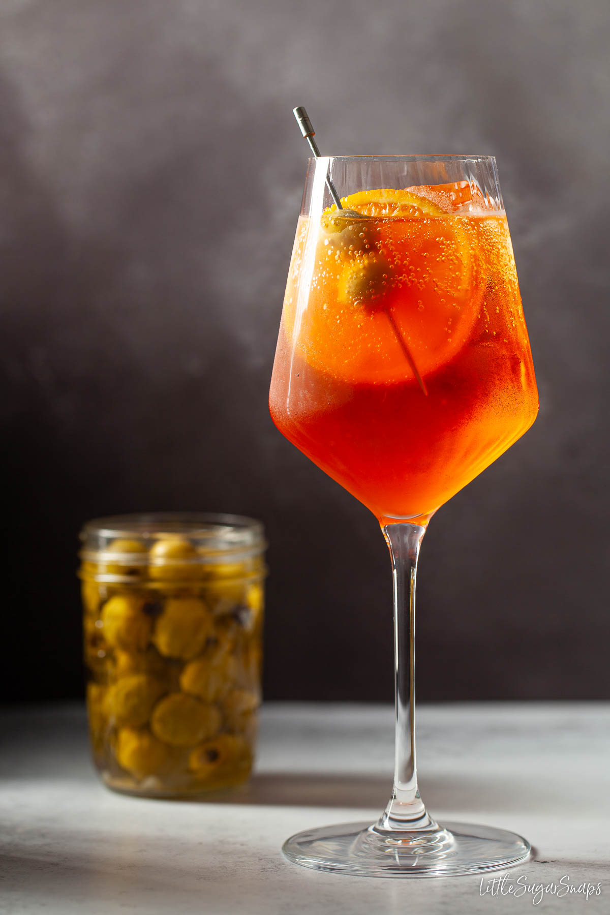Aperol spritz Veneziano with orange and olive garnish and a jar of olives behind.