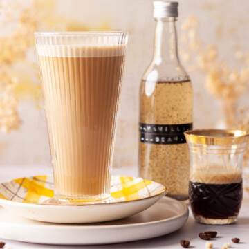 Glass of vanilla bean latte with a bottle of syrup and espresso coffee next to it.