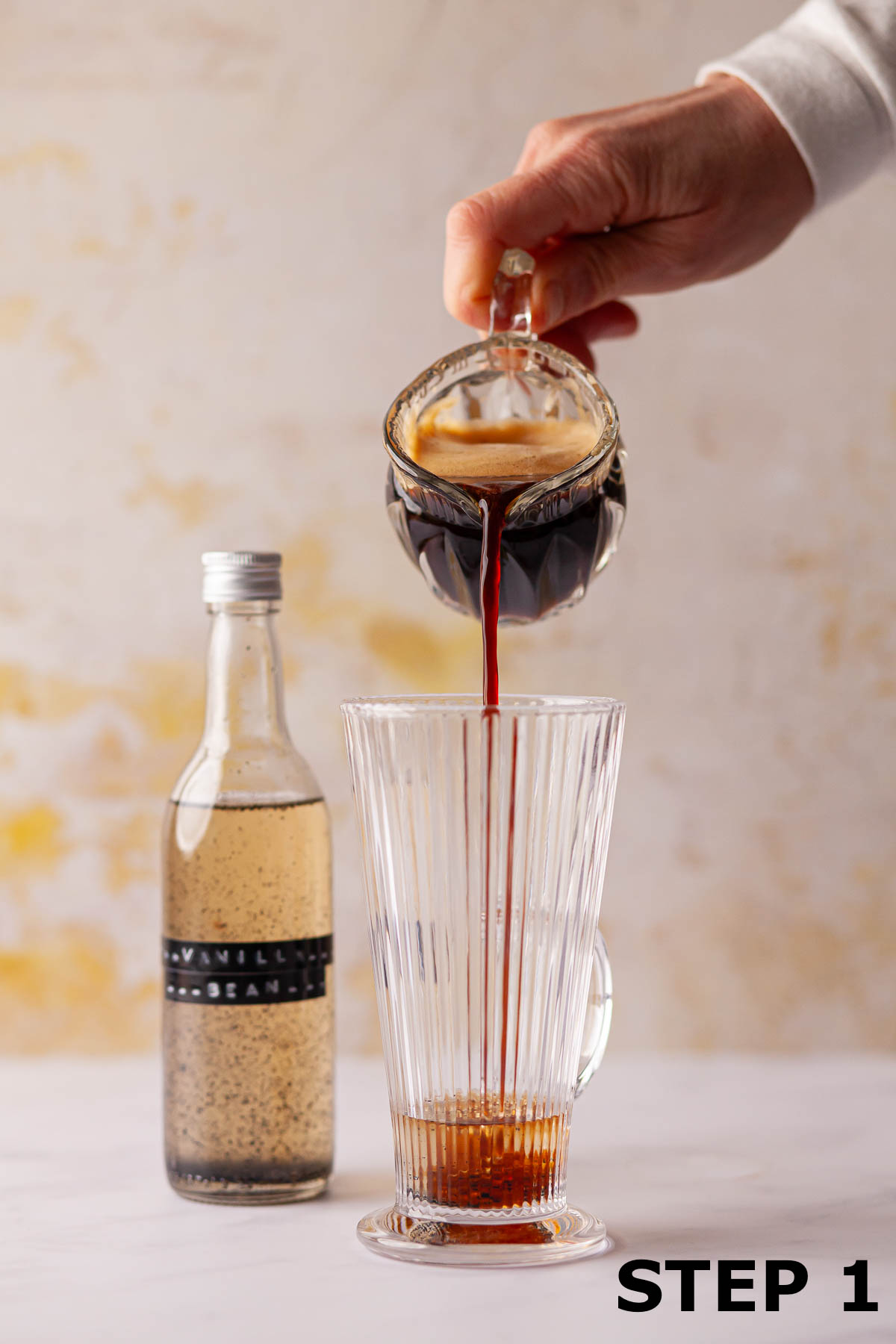 A person pouring coffee into a glass with syrup in it with the syrup bottle in the background.