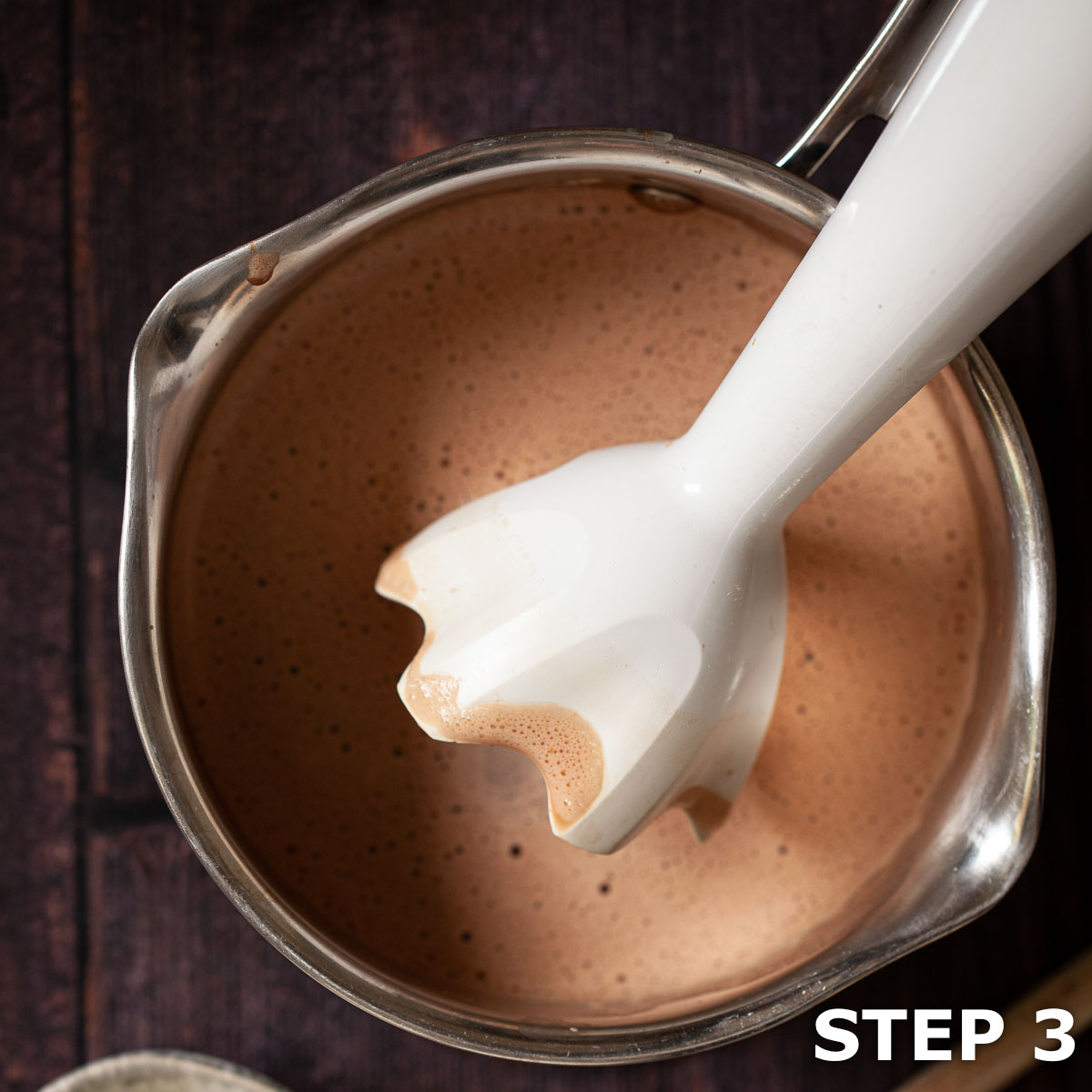 An immersion blender being used to froth hot chocolate in a saucepan.
