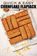 Labelled cornflake flapjack slices on parchment with cornflkaes and oats on the worktop.