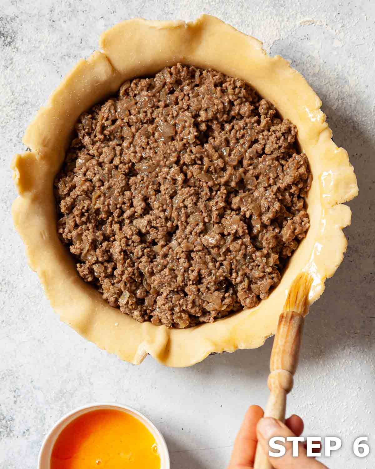Minced beef and onion pie filling and egg wash being brushed around the edge of the pastry case.