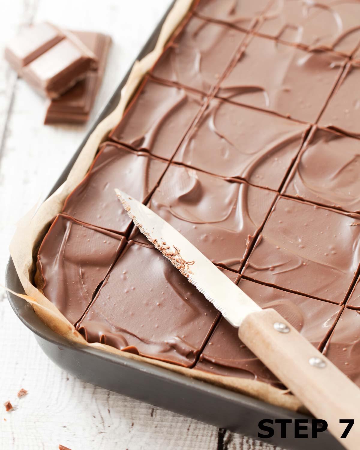 Rice Krispies bars with caramel and chocolate cut into squares in a baking tin with a knife on top.