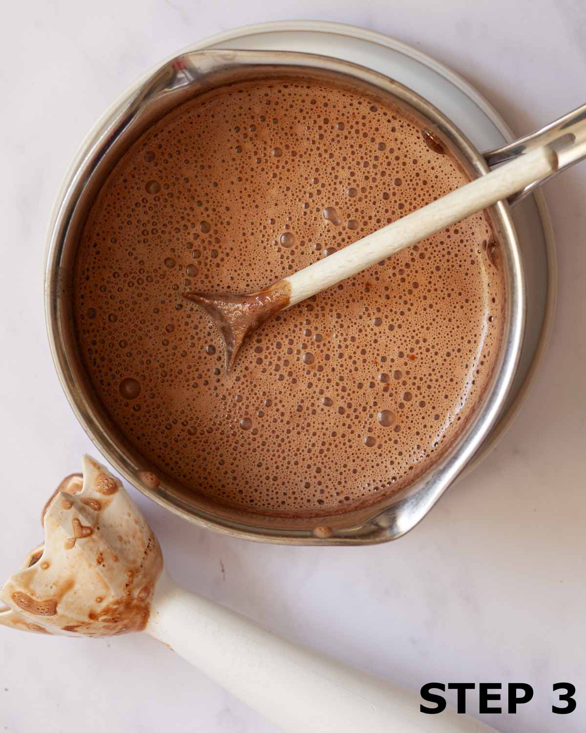 Hot chocolate in a saucepan with an immersion blender on the worktop.