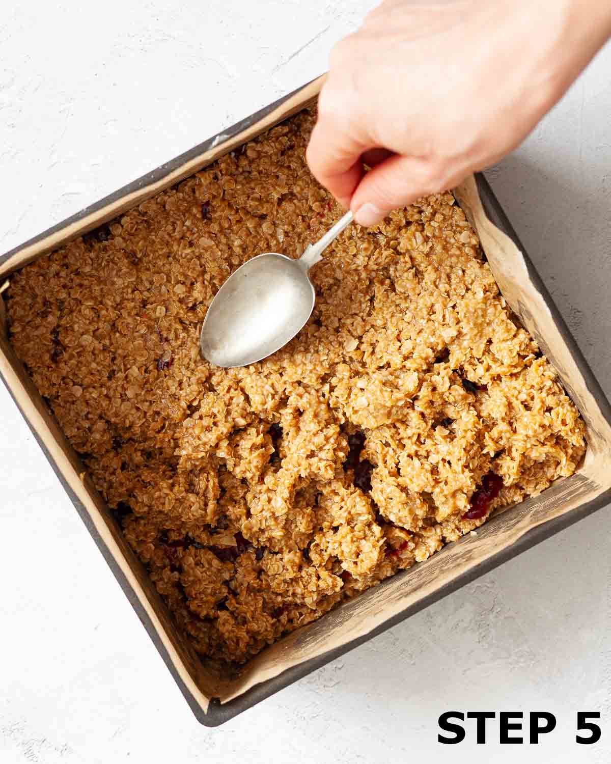 A person pressing peanut butter flapjack mixture into a baking tin using a spoon.