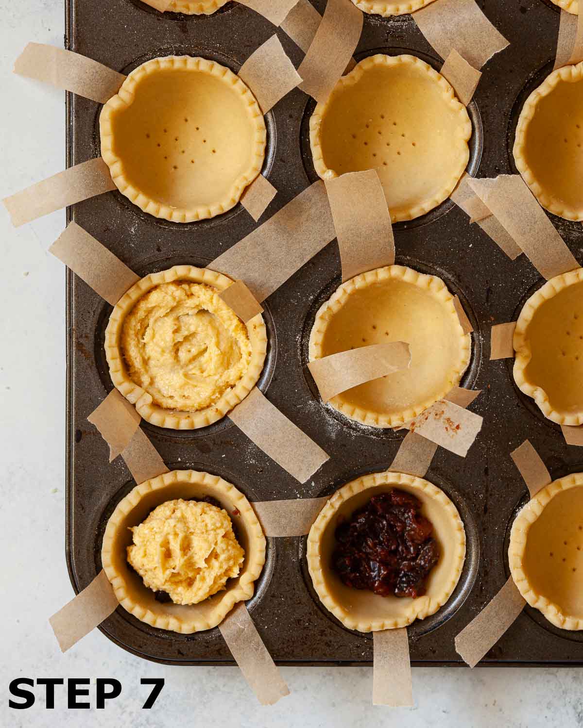 Uncooked pastry shells in a muffin tin being filled with mincemeat and frangipane batter/.
