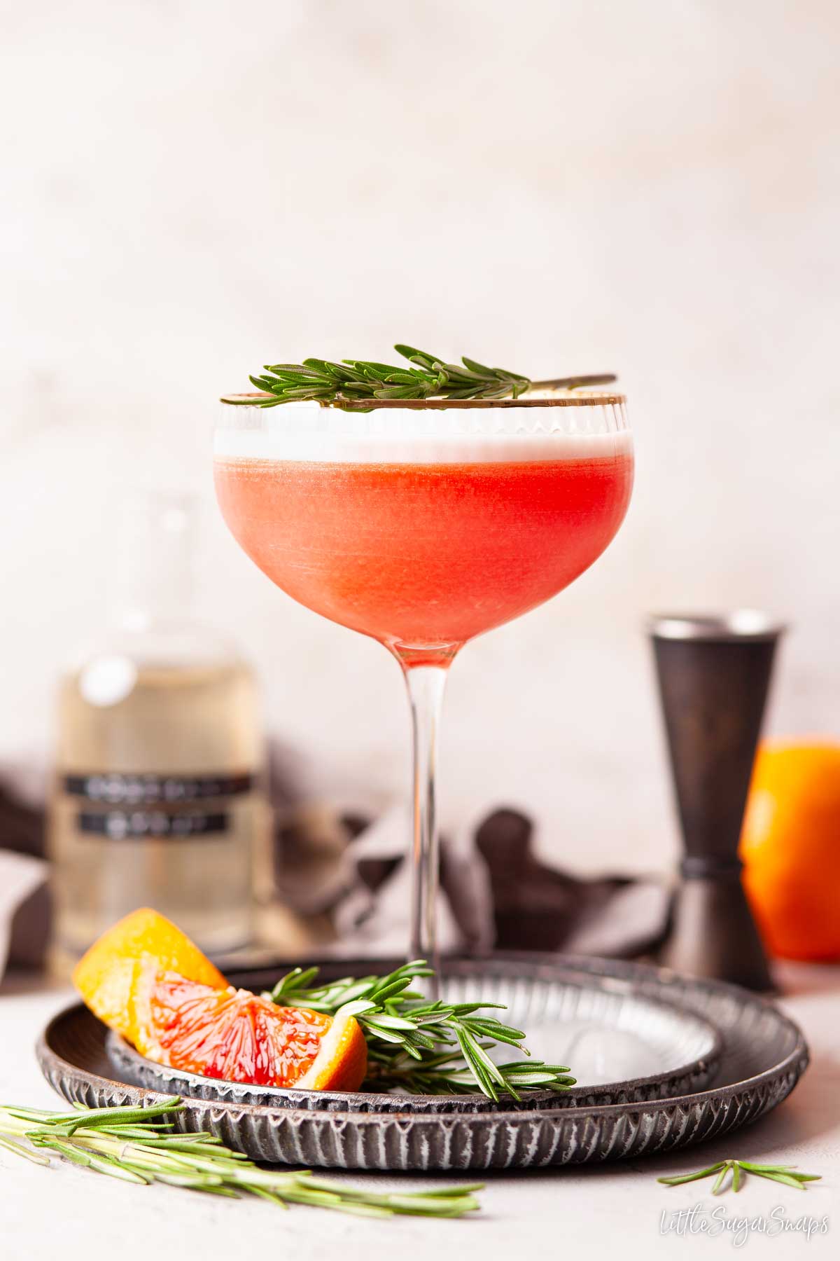 Blood orange sour cocktail with egg white top and rosemary garnish with rosemary and blood orange on a plate.