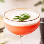 A blood orange cocktail with egg white top and rosemary garnish.