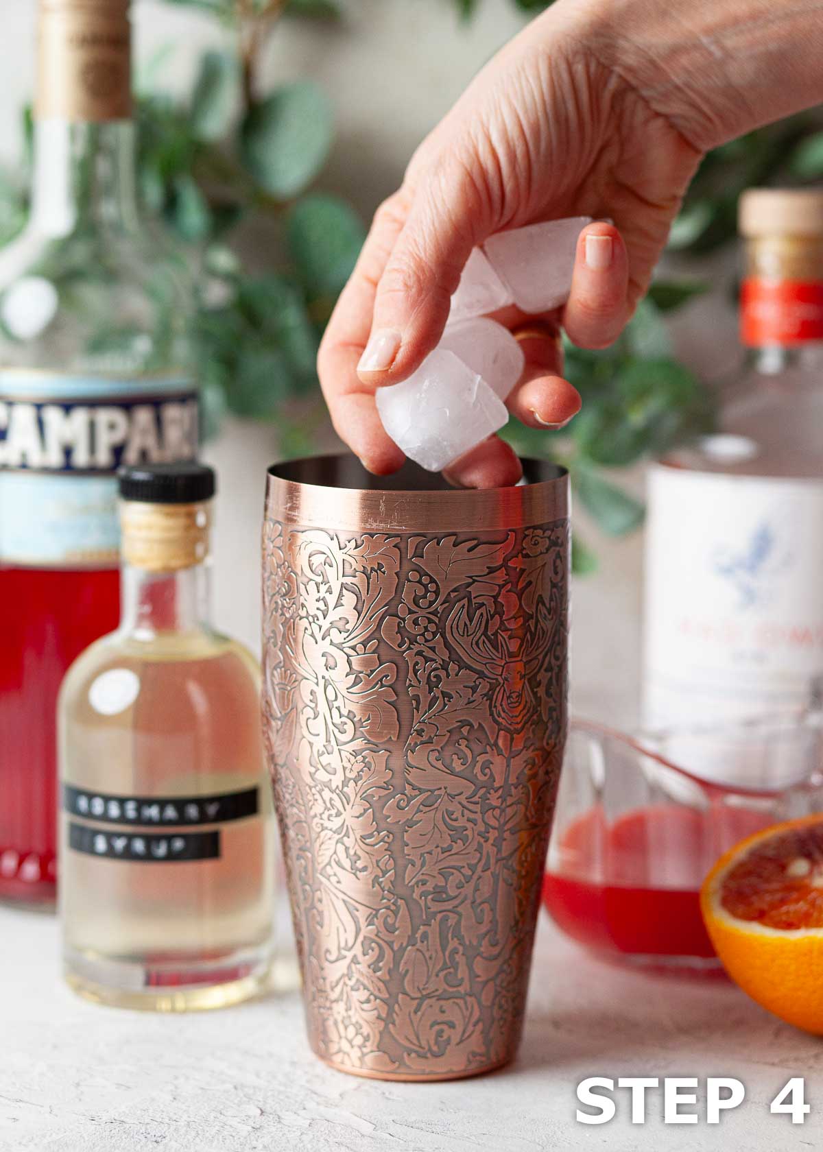 Ice being addd to an embossed copper cocktail shaker with cocktail ingredients in the background.