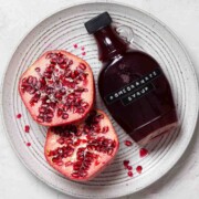 A bottle of pomegranate juice on a plate with a fresh pomegranate fruit cut in half.