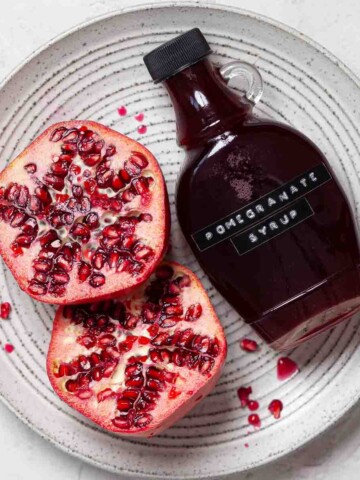 A bottle of pomegranate juice on a plate with a fresh pomegranate fruit cut in half.
