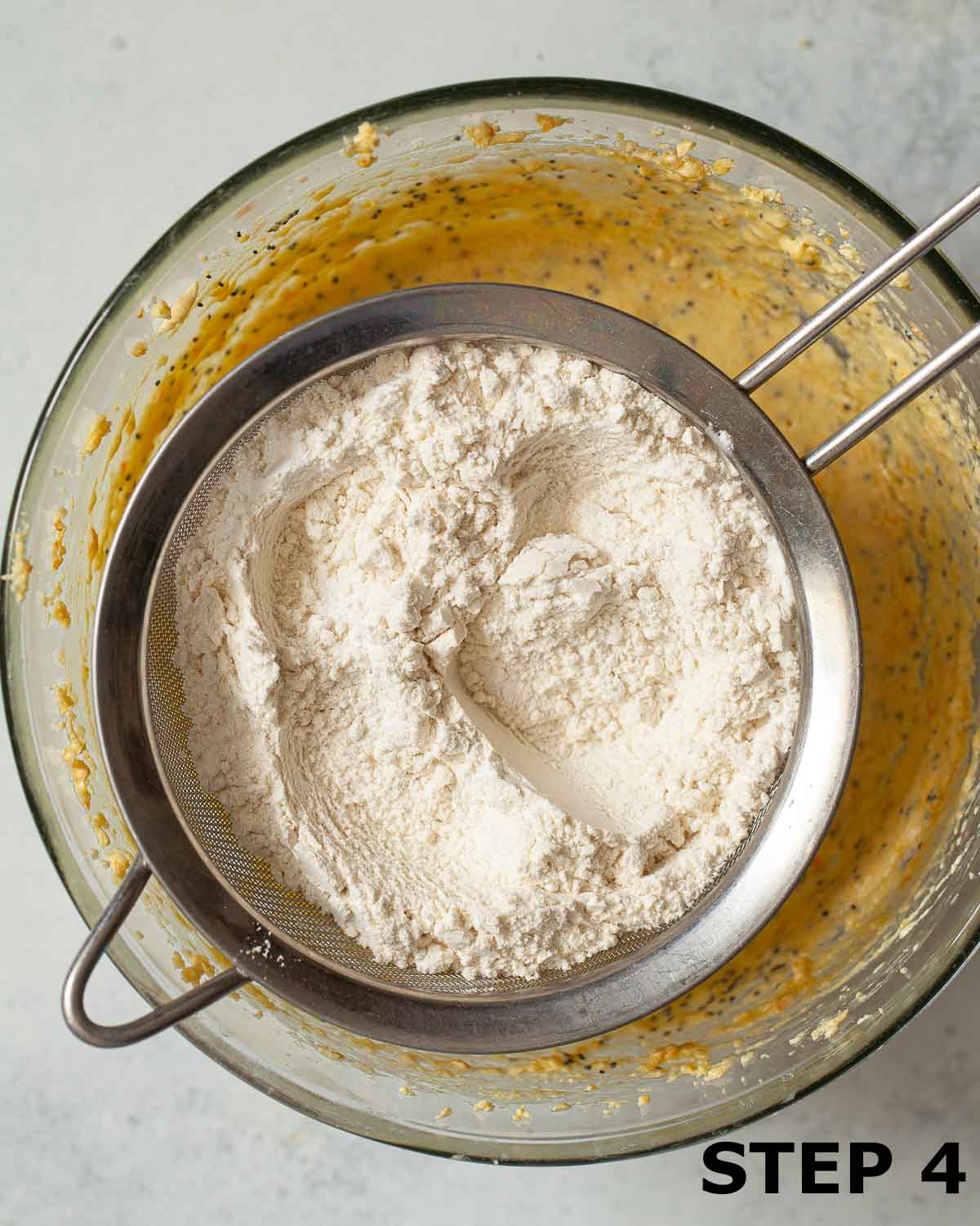 Flour in a sieve suspended over a bowl of cake batter.