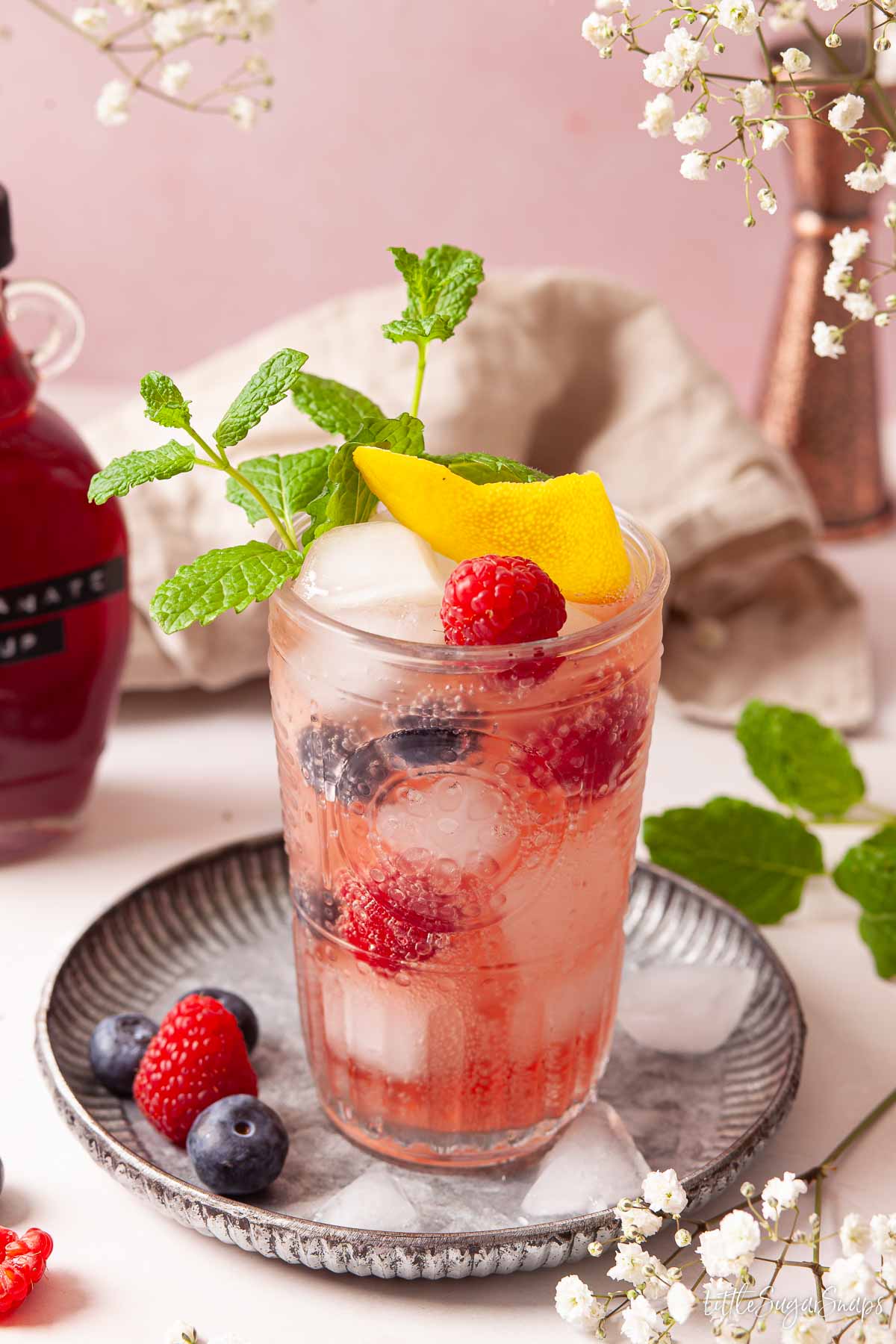 Pomegranate cocktail in a highball glass with lemon, mint and berry garnish.