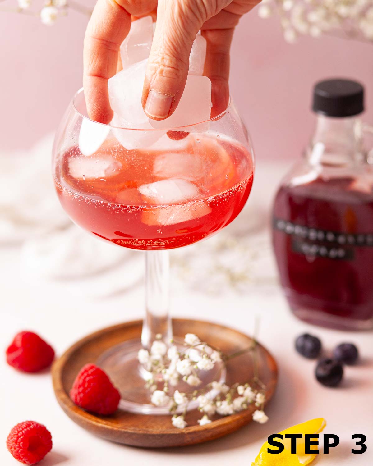 A person adding ice to a pomegranate and lemonade mocktail in a copa glass.