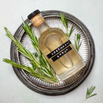 A labelled bottle of rosemary simple syrup on a metal plate with fresh rosemary sprigs.
