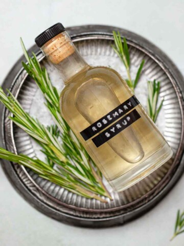 A labelled bottle of rosemary simple syrup on a metal plate with fresh rosemary sprigs.