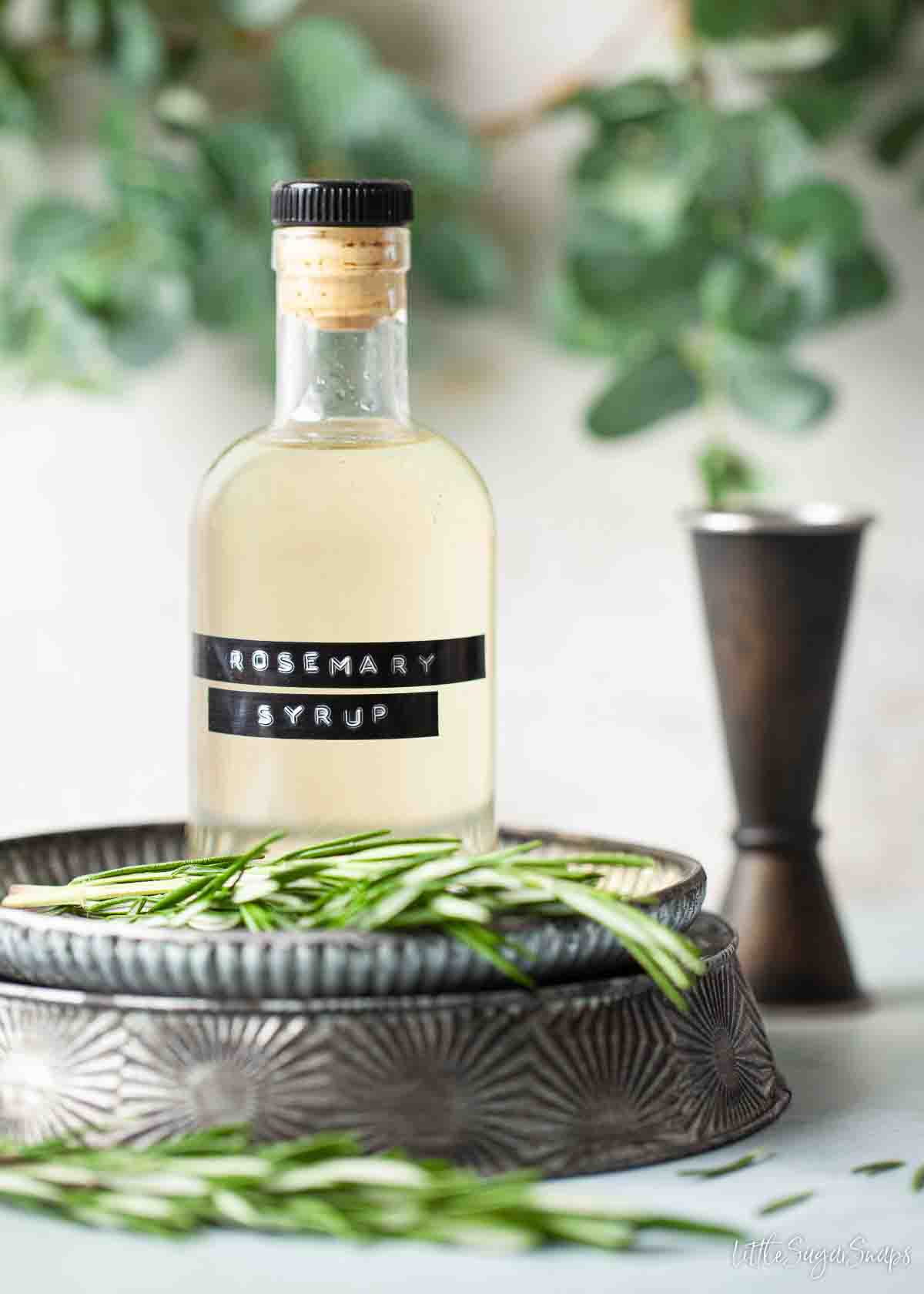 Rosemary syrup for cocktails with a jigger in the background and fresh rosemary on a plate.