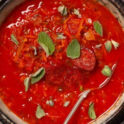 A bowl of carrot and tomato soup garnished with fried chorizo and fresh oregano.