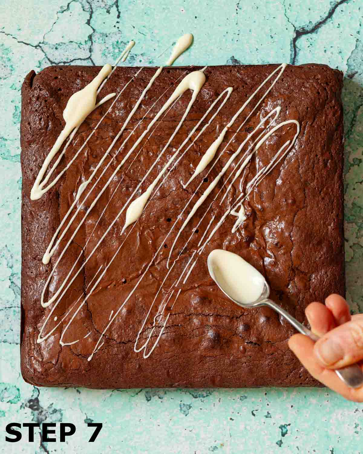 A person drizzling melted white chocolate over a slab of cooked brownies.