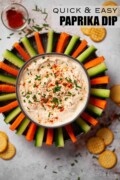 Labelled bowl of paprika soured cream dip with carrot and cucumber sticks surrounding it.