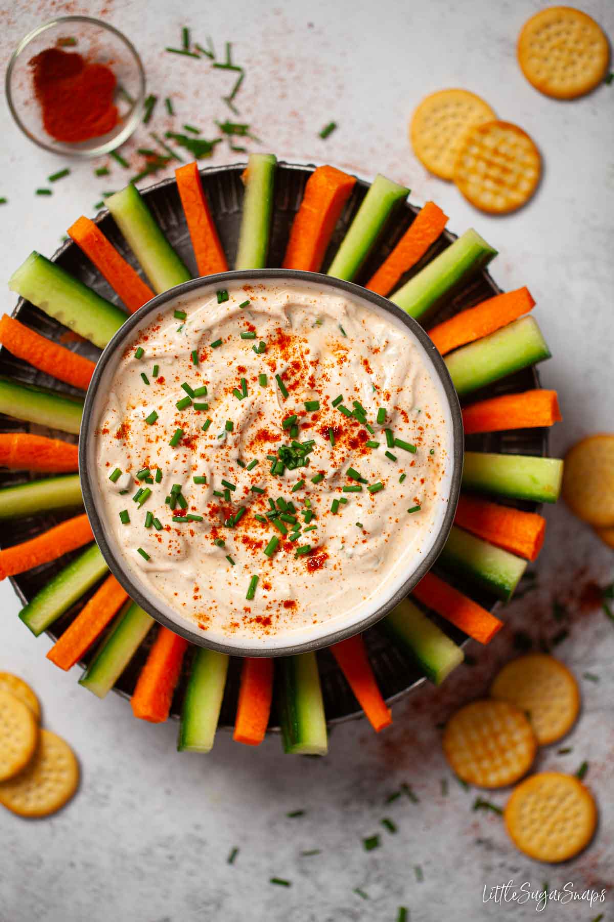 A bowl of paprika soured cream dip with carrot and cucumber sticks surrounding it.