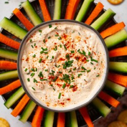 A bowl of paprika soured cream dip with carrot and cucumber sticks surrounding it.
