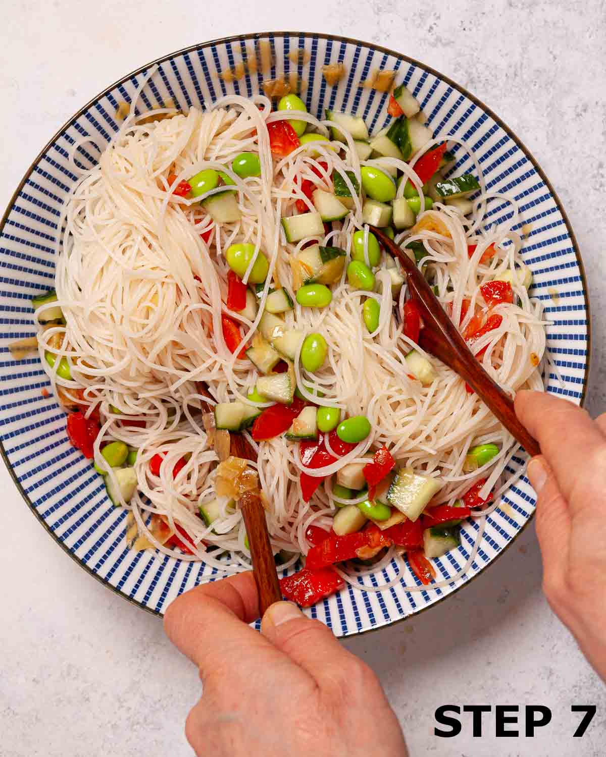 A person mixing ingredients for cucumber tomato rice noodle salad together in a bowl using wooden spoons.