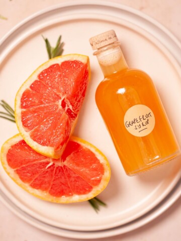 A bottle of grapefruit syrup on a plate with slices of red grapefruit.