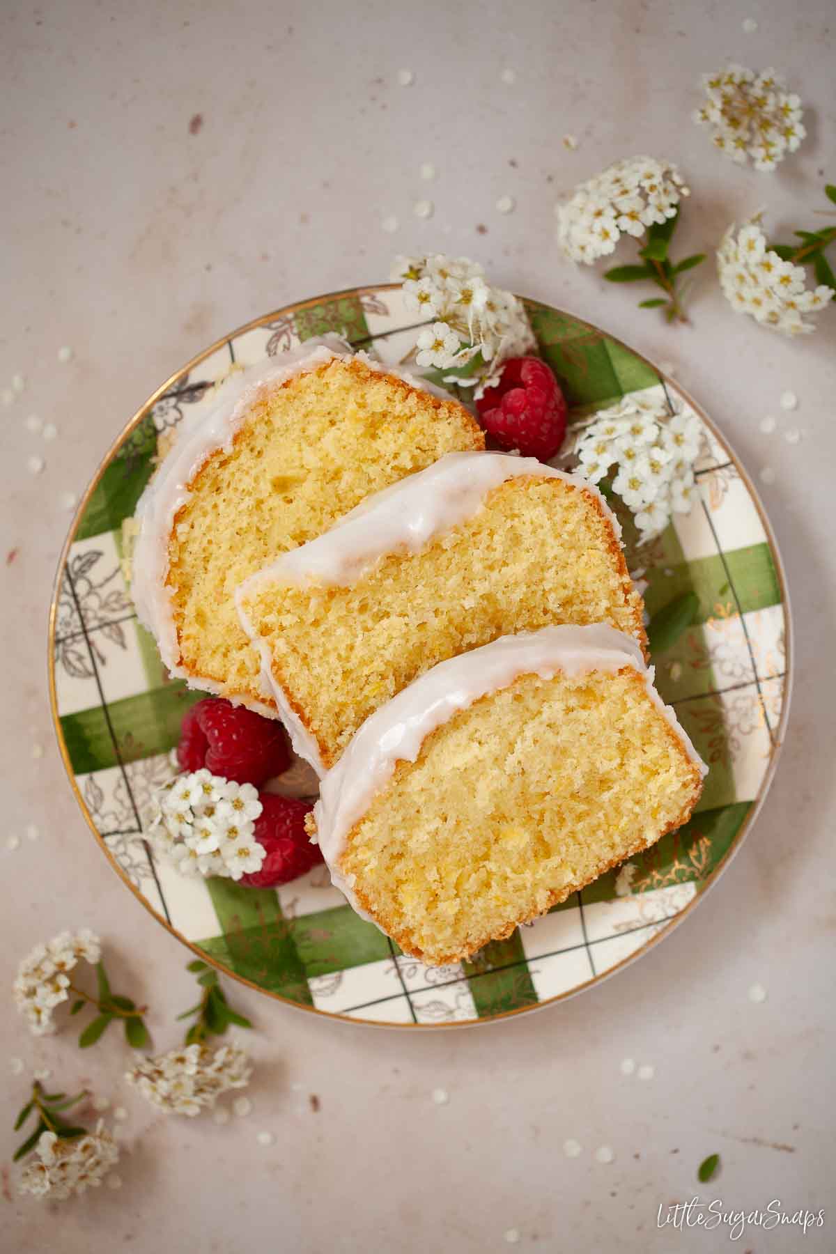 Three slices of iced Leicester sand cake on a plate with flowers and raspberries.