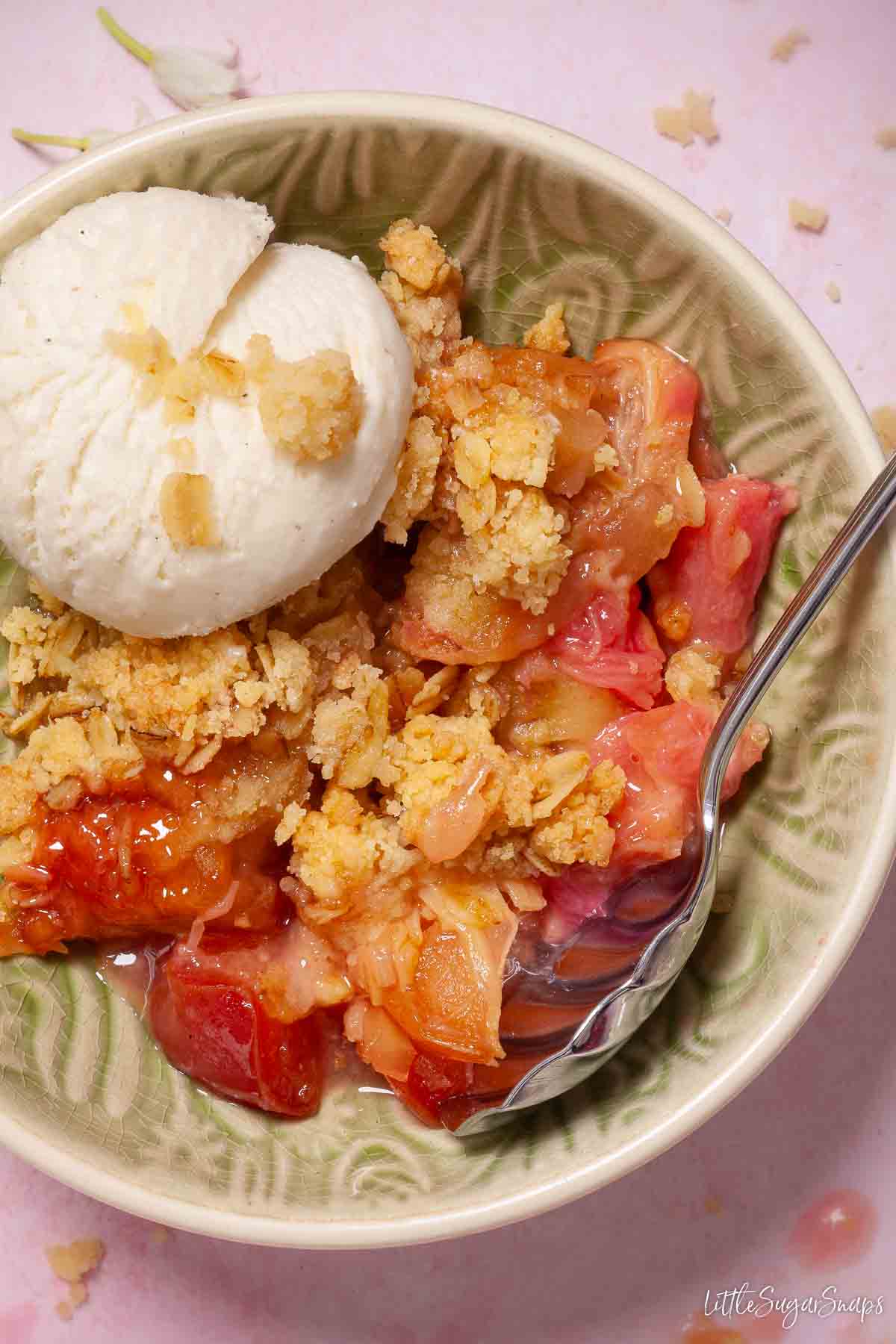 A bowl of apple and rhubarb crumble topped with ice cream with a spoon in the bowl.