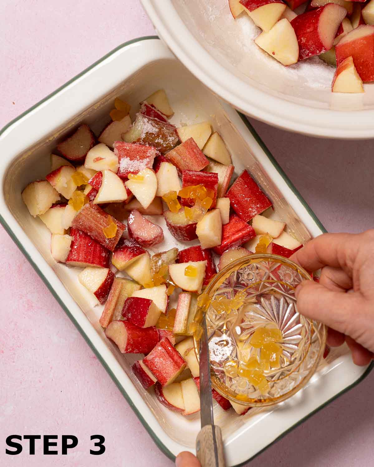 A person scattering chopped stem ginger over apple ands rhubarb in a baking dish.
