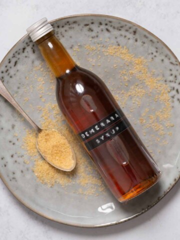 A labelled bottle of Demerara syrup on a plate with a spoon and sugar alongside.
