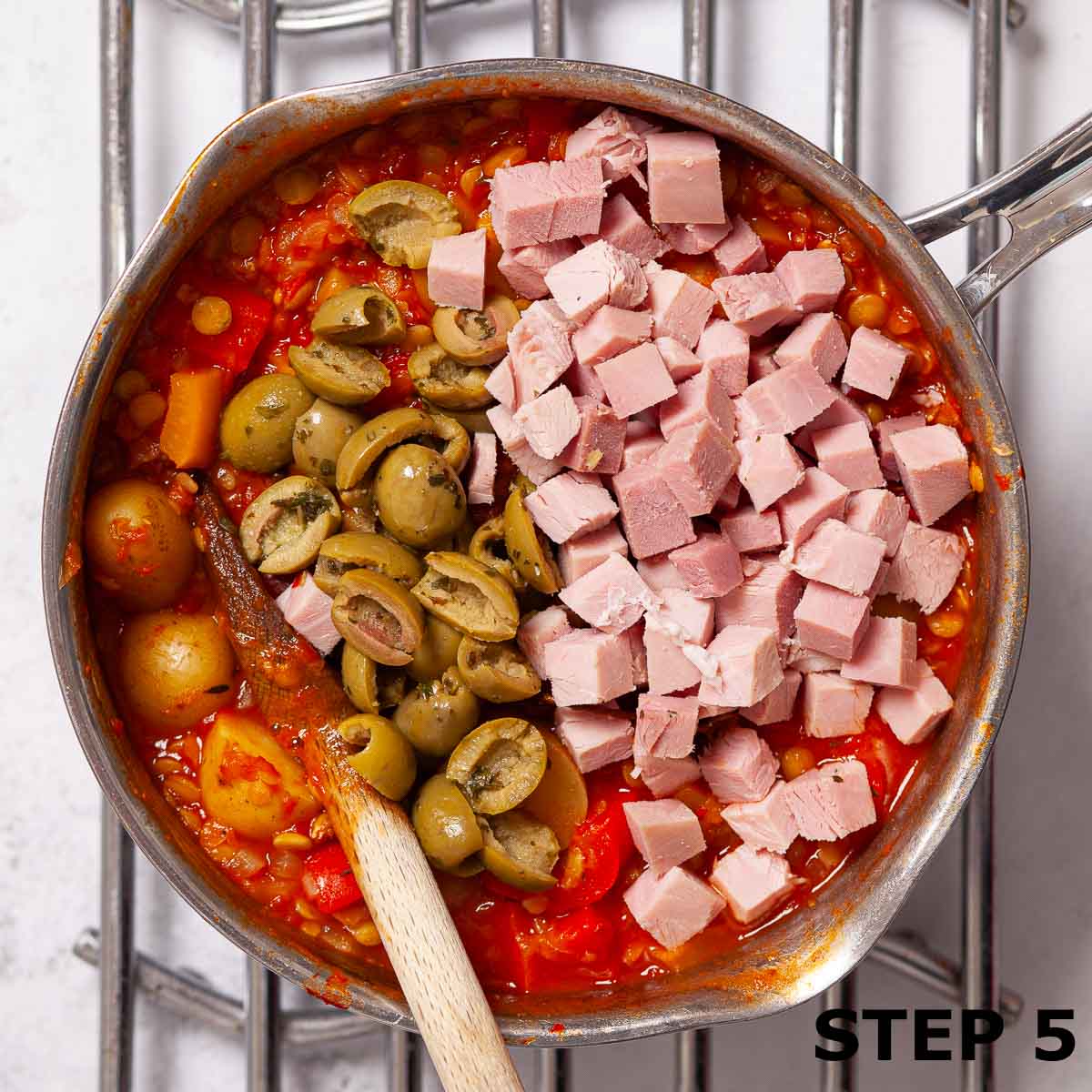 Cubed ham and sliced green olives on top of lentil and potato stew in a saucepan.